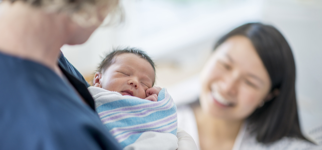 Nurse holds newborn baby wrapped in blanket as mother looks on smiling