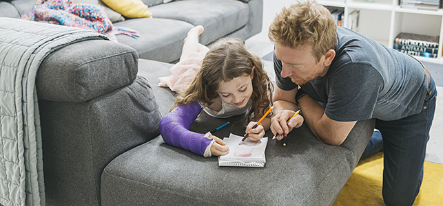 Girl with purple arm cast coloring with her dad on the sofa