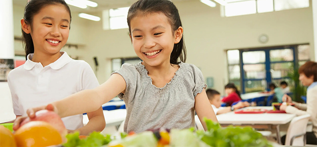 Two smiling girls selecting fruit and veggies in school cafeteria